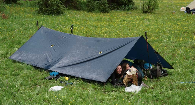 A group of people huddle under a tarp shelter set up in a green flat space 